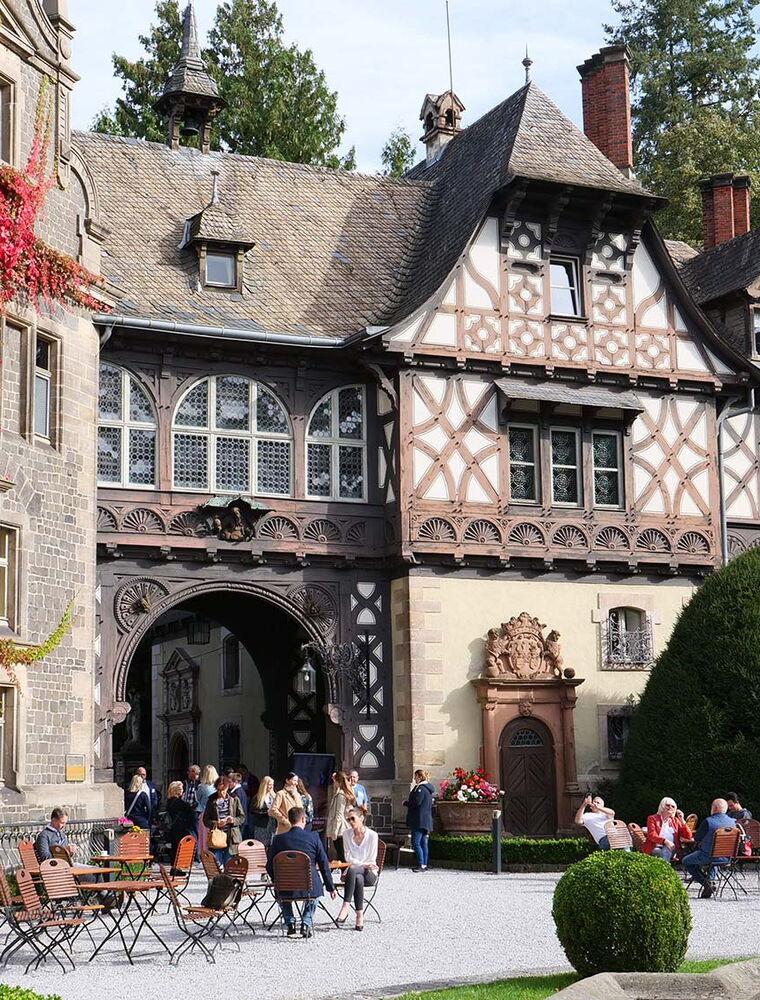 Blick auf das historische Fachwerkschloss Rauischholzhausen bei Sonnenschein. Auf dem Vorplatz sitzen Menschen an Tischen und unterhalten sich.