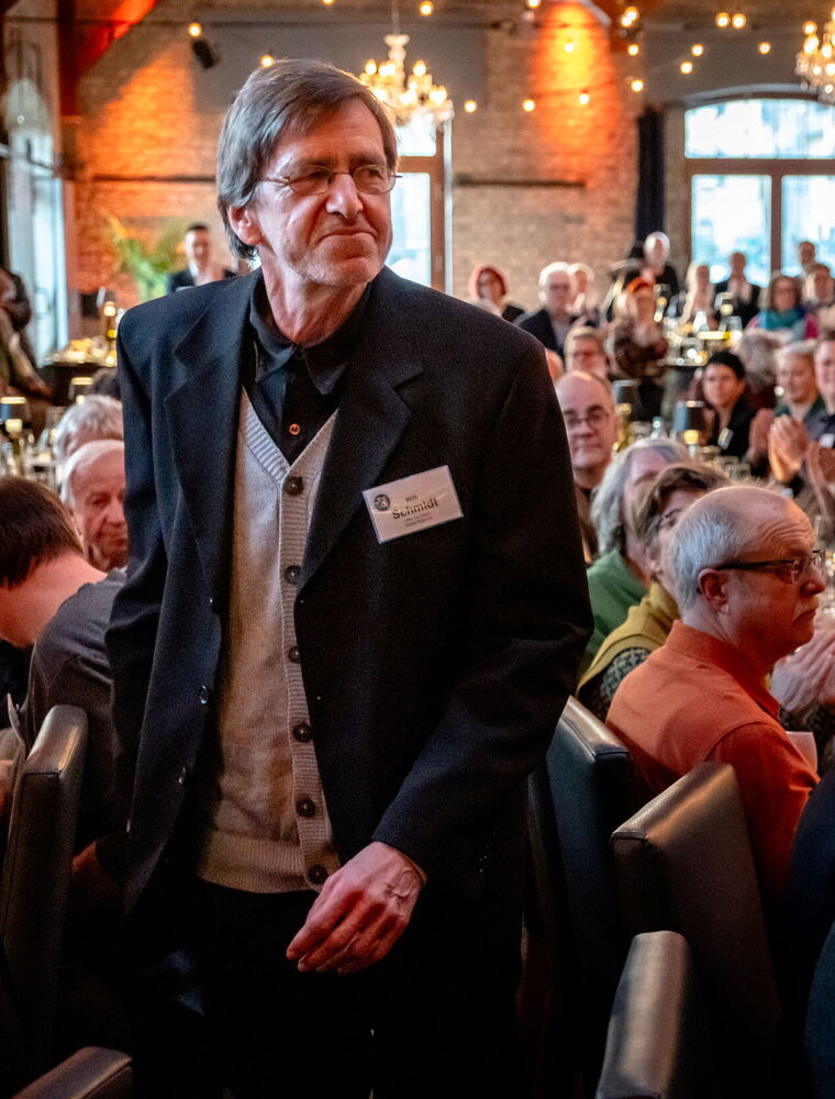 Der Preisträger Willi Schmidt ist auf dem Weg zur Bühne und geht dazu durch eine Reihe Beifall klatschenden Gäste.