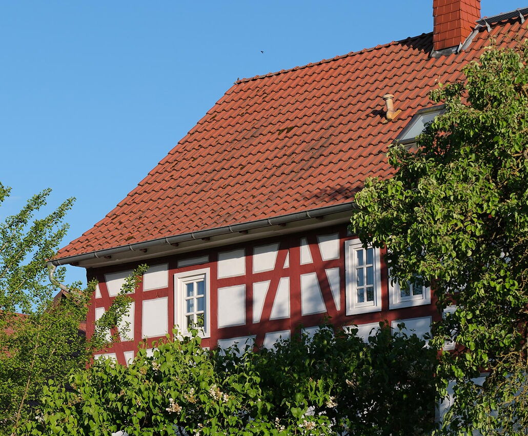 Ein rotes Fachwerkhaus wird vom Sonnenlicht angestrahlt. Man sieht das Haus nur ab dem oberen Geschoss, der unter teil des Bildes wird von einem alten Zaun bestimmt, der mit Blättern bewachsen ist.
