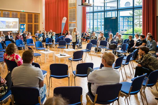 Ein Stuhlkreis mit mehreren Reihen. Auf den Stühlen sitzen Menschen und hören einer Frau zu, die in der Mitte des Kreises steht und mit den anderen spricht.