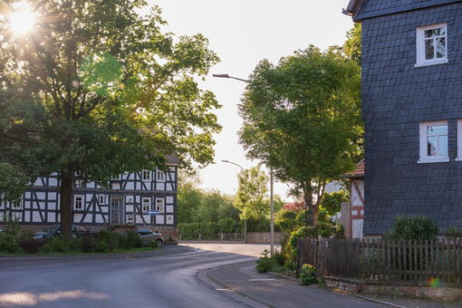 An einer Durchgangsstraße in einem kleinen Ort stehen auf beiden Seiten Fachwerkhäuser. Vor dem linken steht ein Baum, das rechte ist verschiefert. Durch die Blätter und Äste des Baums strahlt die Abendsonne.