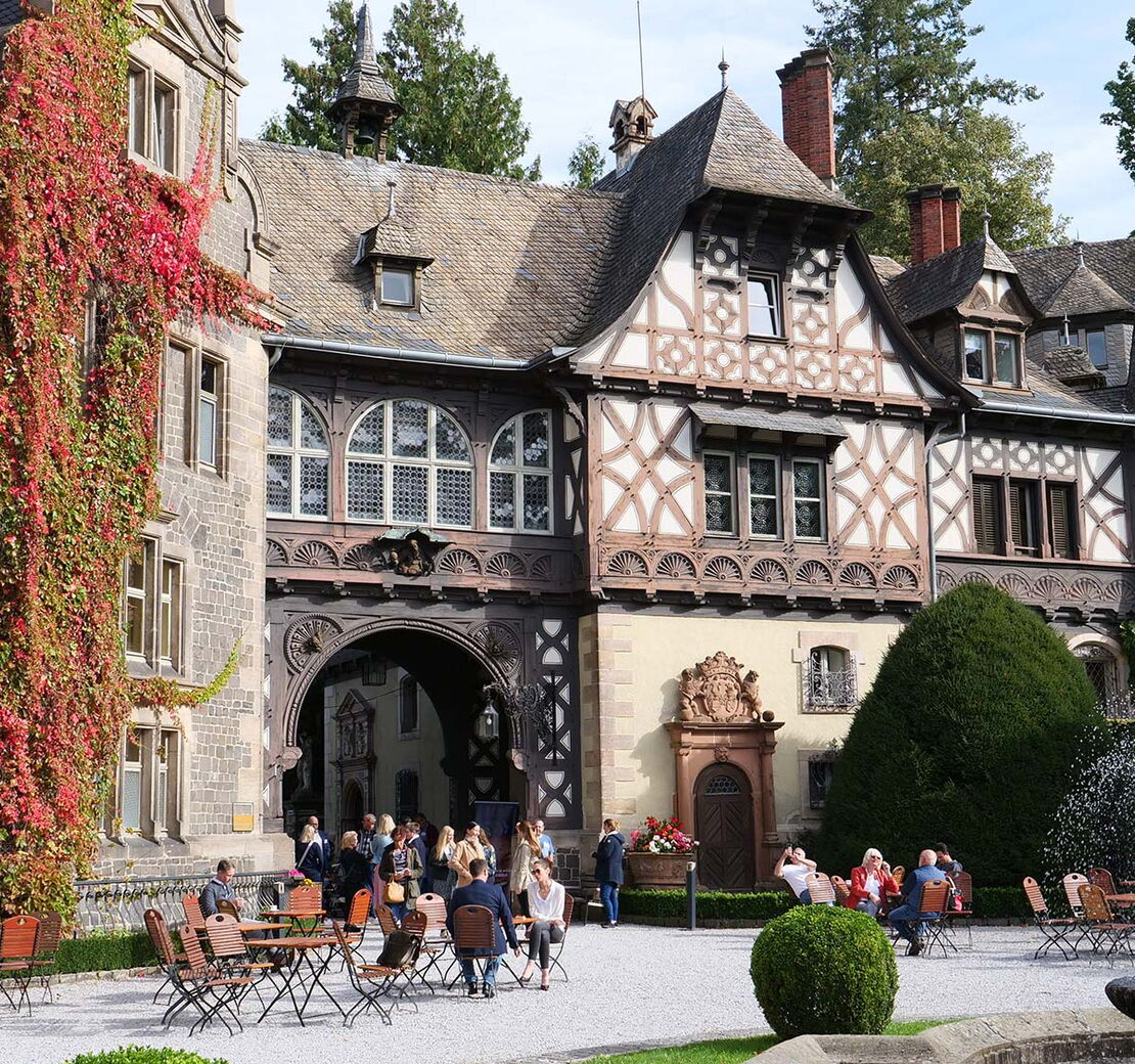 Blick auf das historische Fachwerkschloss Rauischholzhausen bei Sonnenschein. Auf dem Vorplatz sitzen Menschen an Tischen und unterhalten sich.