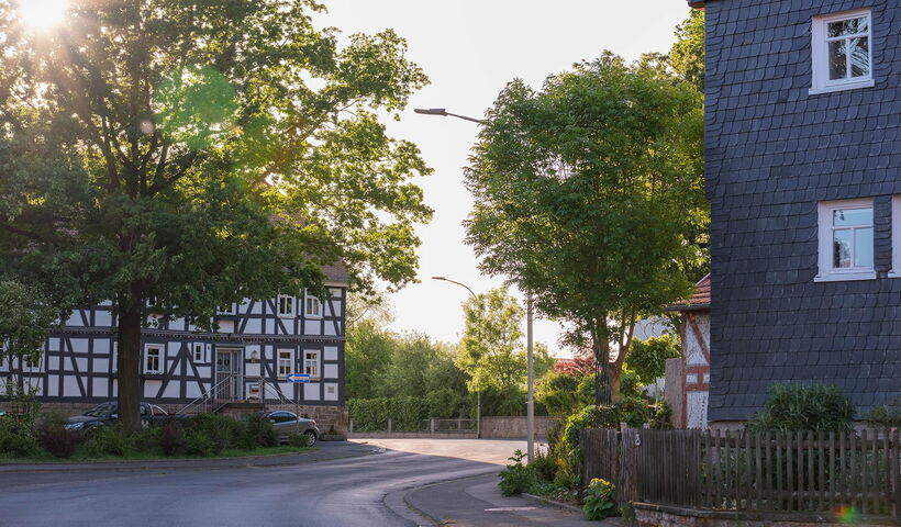 An einer Durchgangsstraße in einem kleinen Ort stehen auf beiden Seiten Fachwerkhäuser. Vor dem linken steht ein Baum, das rechte ist verschiefert. Durch die Blätter und Äste des Baums strahlt die Abendsonne.